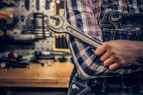 Mechaniker mit Schraubenschlüssel in Werkstatt Ein Mechaniker in kariertem Hemd und Latzhose hält einen großen Schraubenschlüssel vor sich.